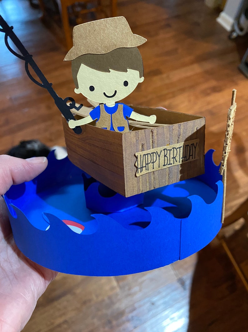 Puede incluir: Una tarjeta de cumplea&ntilde;os de papel con un pescador de dibujos animados en un bote con una ca&ntilde;a de pescar y las palabras "Feliz cumplea&ntilde;os" en el lateral. El bote est&aacute; sentado sobre olas azules.