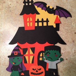 May include: A Halloween-themed cake topper featuring a black and orange house with a witch, a Frankenstein, and a jack-o'-lantern. The house has a red roof and yellow windows. The witch is wearing a purple hat and green dress. The Frankenstein is wearing a gray shirt and black pants. The jack-o'-lantern is smiling.