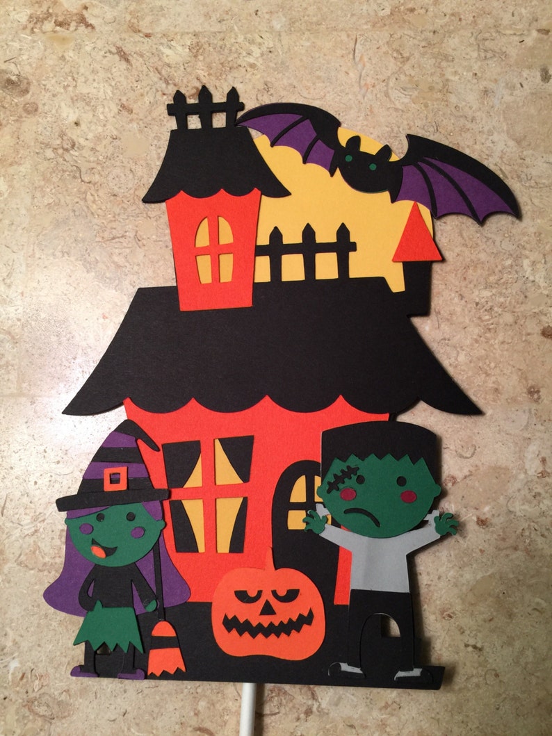 May include: A Halloween-themed cake topper featuring a black and orange house with a witch, a Frankenstein, and a jack-o'-lantern. The house has a red roof and yellow windows. The witch is wearing a purple hat and green dress. The Frankenstein is wearing a gray shirt and black pants. The jack-o'-lantern is smiling.