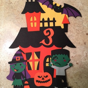 May include: A Halloween-themed cake topper featuring a black and orange house with a number 3, a witch, a Frankenstein, and a jack-o-lantern.