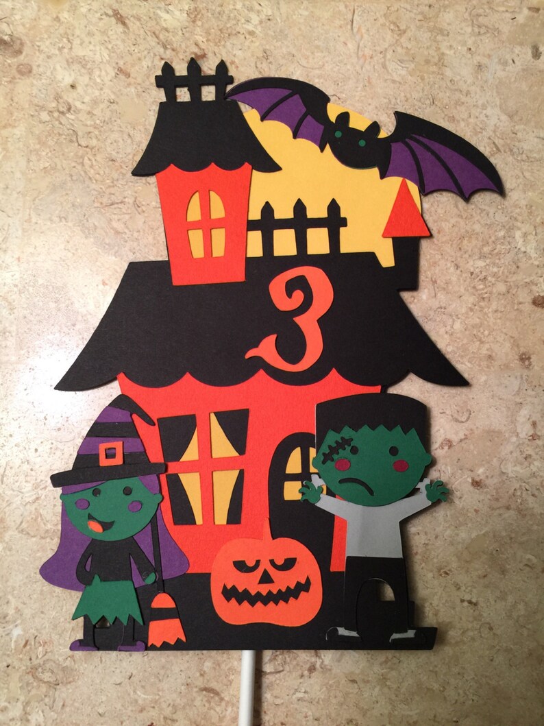 May include: A Halloween-themed cake topper featuring a black and orange house with a number 3, a witch, a Frankenstein, and a jack-o-lantern.