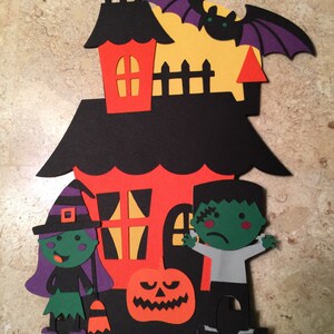 May include: A Halloween-themed cake topper featuring a black house with orange windows, a witch, a Frankenstein, a pumpkin, and a bat. The house has a yellow moon behind it.
