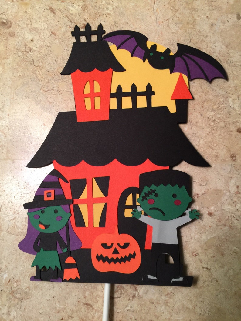 May include: A Halloween-themed cake topper featuring a black house with orange windows, a witch, a Frankenstein, a pumpkin, and a bat. The house has a yellow moon behind it.