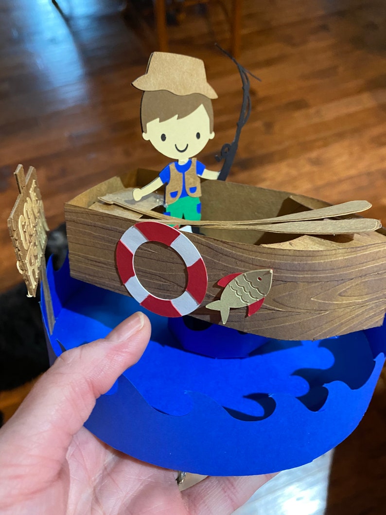 Puede incluir: Una figurita de papel de un ni&ntilde;o con un sombrero marr&oacute;n y una camisa azul que sostiene una ca&ntilde;a de pescar en un barco marr&oacute;n con un salvavidas rojo y blanco y un pez marr&oacute;n. El barco est&aacute; sentado sobre una ola de papel azul.