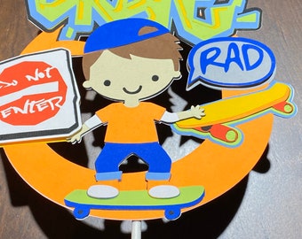 Puede incluir: Un adorno para pastel con un niño de dibujos animados que lleva un sombrero azul y una camisa naranja, haciendo skateboard con una tabla amarilla y naranja. El fondo presenta una palabra de graffiti verde y amarilla "Skate" y un globo de diálogo con la palabra "RAD" en letras azules.
