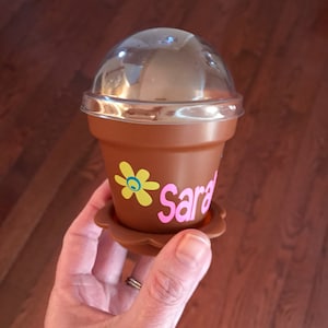May include: A brown plastic planter with a clear plastic lid. The planter has a yellow flower and the name "Sara" written in pink letters.