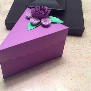 Puede incluir: Una caja de regalo triangular de color lila con un borde festoneado. Está adornada con flores de papel en tonos morados y verdes. El arreglo floral incluye una rosa grande y varios capullos más pequeños.