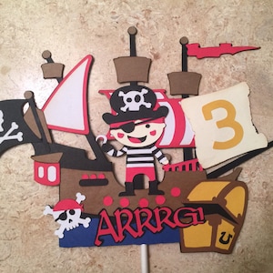 May include: A pirate-themed cake topper featuring a cartoon pirate holding a sword, a ship with a skull and crossbones flag, a treasure chest, and the number 3. The text "ARRRG!" is also included.