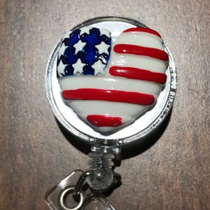 Puede incluir: Un portagafete retráctil plateado con un dije en forma de corazón que presenta la bandera estadounidense. El dije está pintado con estrellas y rayas rojas, blancas y azules.