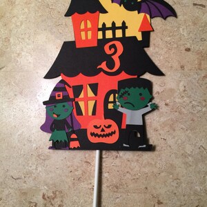 May include: A black, orange, and green Halloween cake topper with a haunted house, a witch, a Frankenstein, a pumpkin, and the number 3.