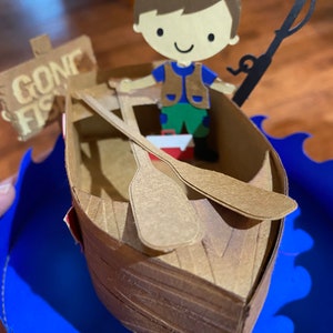 Puede incluir: Una figurita de papel de un ni&ntilde;o con un sombrero marr&oacute;n y un chaleco, sosteniendo una ca&ntilde;a de pescar y sentado en un bote marr&oacute;n con un letrero que dice "Gone Fishin'" en el lateral. El bote est&aacute; sobre una ola azul.
