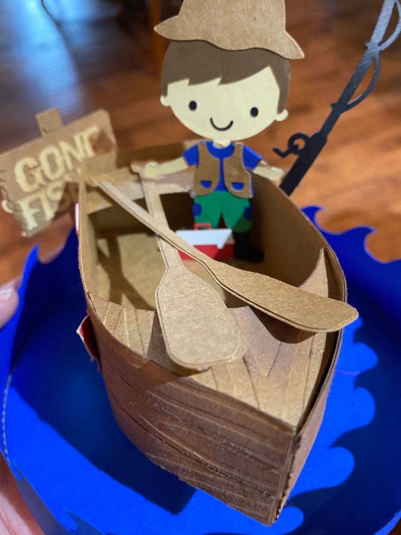 Puede incluir: Una figurita de papel de un ni&ntilde;o con un sombrero marr&oacute;n y un chaleco, sosteniendo una ca&ntilde;a de pescar y sentado en un bote marr&oacute;n con un letrero que dice "Gone Fishin'" en el lateral. El bote est&aacute; sobre una ola azul.