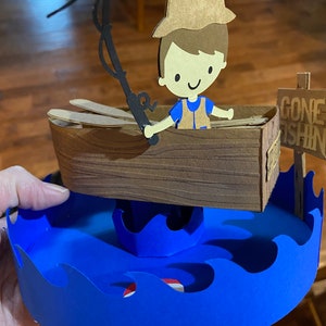 Puede incluir: Una figurita de papel de un ni&ntilde;o con un sombrero marr&oacute;n y una camisa azul que sostiene una ca&ntilde;a de pescar en un bote marr&oacute;n. El bote est&aacute; sentado sobre una base de papel azul con olas. Un letrero dice "Gone Fishin'" en texto negro.