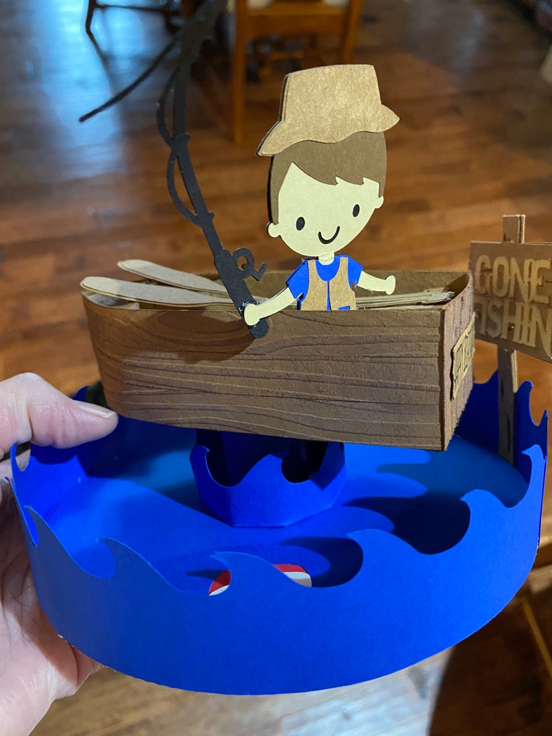 Puede incluir: Una figurita de papel de un ni&ntilde;o con un sombrero marr&oacute;n y una camisa azul que sostiene una ca&ntilde;a de pescar en un bote marr&oacute;n. El bote est&aacute; sentado sobre una base de papel azul con olas. Un letrero dice "Gone Fishin'" en texto negro.