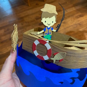 Puede incluir: Una manualidad de papel de un ni&ntilde;o con un sombrero marr&oacute;n y un chaleco verde pescando desde un barco marr&oacute;n con un salvavidas rojo y blanco. El barco est&aacute; encima de una ola azul.