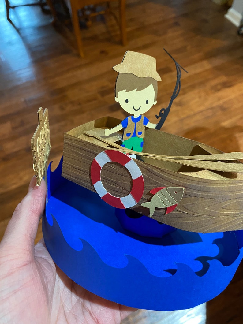 Puede incluir: Una manualidad de papel de un ni&ntilde;o con un sombrero marr&oacute;n y un chaleco verde pescando desde un barco marr&oacute;n con un salvavidas rojo y blanco. El barco est&aacute; encima de una ola azul.