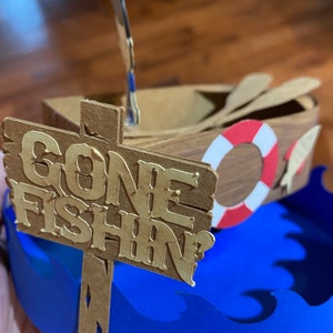 Puede incluir: Un barco de madera con un salvavidas y remos, sentado sobre olas azules con un letrero que dice "Gone Fishin".
