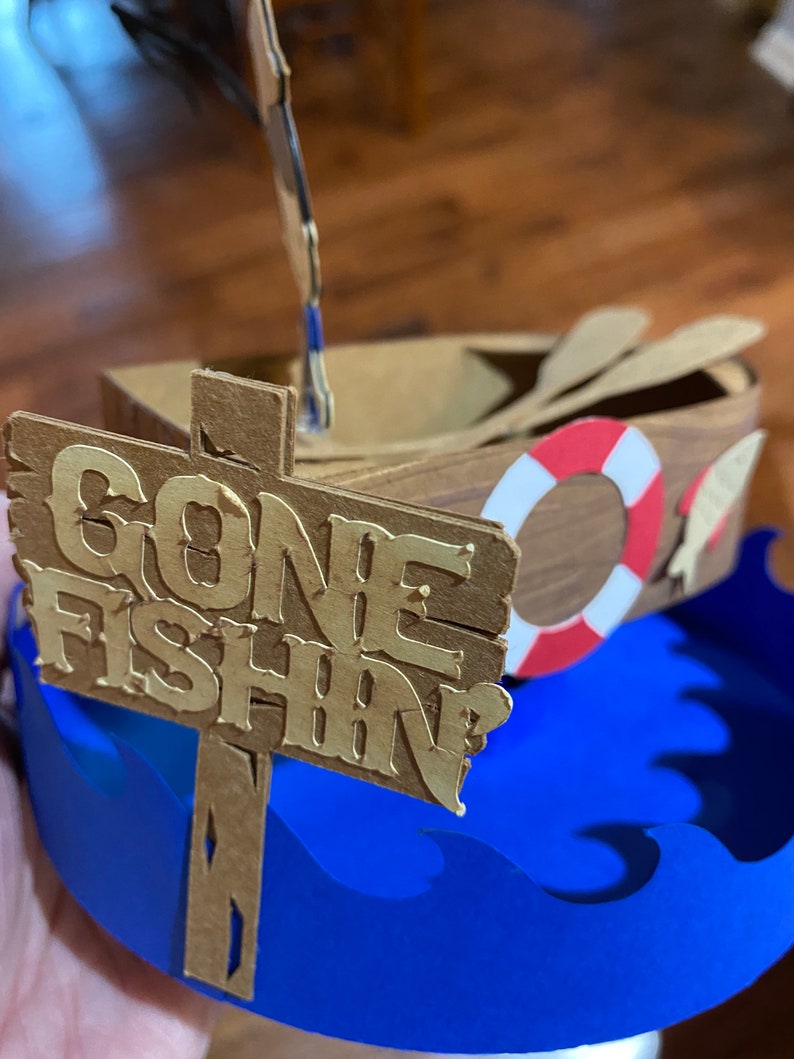 Puede incluir: Un barco de madera con un salvavidas y remos, sentado sobre olas azules con un letrero que dice "Gone Fishin".