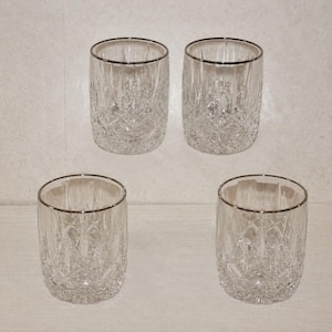May include: Four clear, cut-glass tumblers with silver rims. The glasses have a detailed, crisscross pattern. The tumblers are arranged in a square formation, perfect for serving beverages.