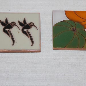 May include: Two square ceramic tiles. The left tile features two black hummingbirds on a cream background. The right tile displays a green pumpkin and an orange pumpkin on a white background. Both tiles have a visible terracotta-colored edge.