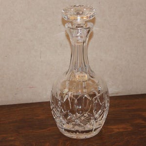May include: A clear crystal decanter with a stopper. The decanter has a bulbous base with a starburst pattern and a slender neck. The stopper is also crystal and has a decorative design. The decanter is on a wooden surface.