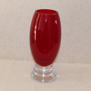 May include: A vibrant red glass vase with a clear, textured base. The vase has a smooth, oval shape and stands approximately 8 inches tall. The base is a series of concentric circles.