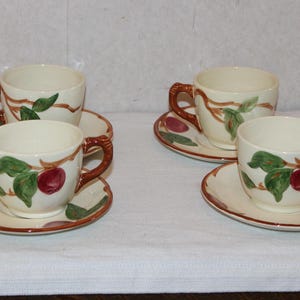 May include: Four cream-colored teacups and saucers. Each cup and saucer features a hand-painted design of red apples, green leaves, and brown branches. The cups have brown handles and a matching brown rim.
