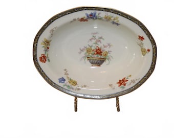 May include: An oval porcelain bowl with a white base, featuring a central floral arrangement in a basket and smaller floral clusters around the rim. The edge is decorated with a dark blue and gold patterned border.