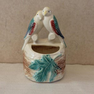 May include: A ceramic wall pocket featuring two white birds with blue and red accents perched above a small opening. The base is decorated with a brown and teal leaf design, creating a nature-inspired aesthetic.