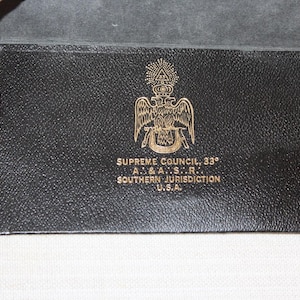 May include: A black leather wallet featuring a gold embossed double-headed eagle emblem above the text SUPREME COUNCIL, 33°, A. & A.S.R., SOUTHERN JURISDICTION, U.S.A. The rectangular accessory has a textured surface and visible black stitching.