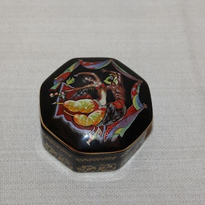 May include: Black, octagonal trinket box with a detailed lid illustration. The lid features a colorful scene with figures and fruit. The box has gold trim and decorative patterns around the base. A unique decorative item.