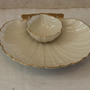 May include: Cream-colored, shell-shaped serving dish with a smaller, matching dish in the center. The edges are trimmed with gold. The dish has a ribbed design, and is likely used for serving appetizers or snacks.
