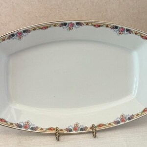 May include: A rectangular white porcelain serving platter with rounded corners, featuring a decorative border of small pink roses, blue flowers, and green leaves, accented by a thin gold trim along the outer edge of the rim.