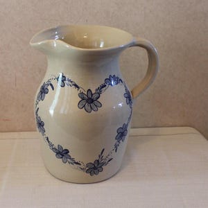 May include: A cream-colored ceramic pitcher with a blue and white floral design in the shape of a heart. The pitcher has a handle and a spout.