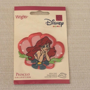May include: Ariel iron-on patch from the Disney Princess Collection. The patch features Ariel with red hair, resting her chin on her hands, surrounded by a pink flower. The packaging includes the Disney Home logo and the Wrights brand.