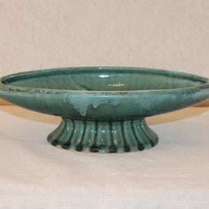 May include: A teal ceramic pedestal bowl with a wide, oval shape. The bowl has a textured, glazed finish and sits on a fluted base. The color is a muted teal with darker accents.
