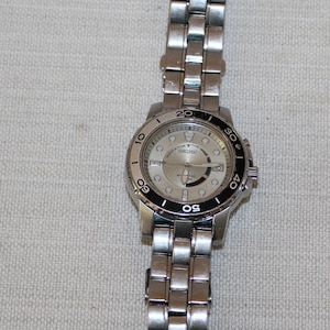 May include: Silver-toned wristwatch with a stainless steel band. The watch face is light-colored with black markings and a black bezel. The watch has a round face and a metal band with rectangular links.