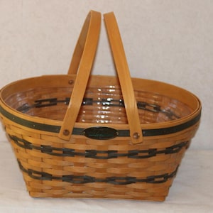 May include: Woven basket with a light brown finish and a dark green trim. The basket has a rectangular base and an oval top, with two wooden handles. A small, dark green oval emblem is on the front.