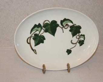 May include: An oval-shaped white ceramic serving platter featuring a central illustration of green ivy leaves and brown vines. The platter is supported by two small, textured gold-toned metal stands positioned at the bottom edge of the dish.