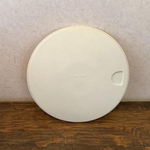 May include: Off-white, round lid with a handle cutout. The lid has the word "Longaberger" printed on it. The lid is sitting on a wooden surface.