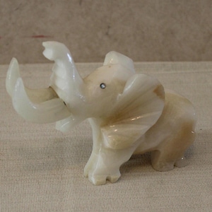 May include: A small, carved ivory-colored elephant figurine with its trunk raised. The elephant has detailed features, including visible ears and tusks. The figurine is made of a stone-like material and sits on a neutral surface.
