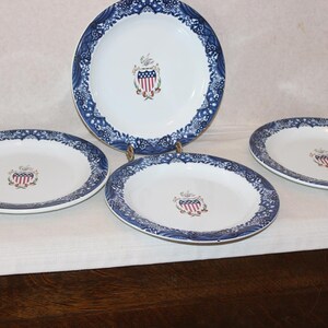 May include: A set of four white ceramic plates with a blue floral border. Each plate features a central emblem with red, white, and blue elements. The plates vary in size, with one larger plate and three smaller ones.