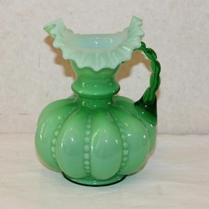May include: A green glass pitcher with a ruffled rim and a twisted handle. The pitcher has a bulbous body with vertical ridges and small, raised dots. The handle is a darker shade of green.
