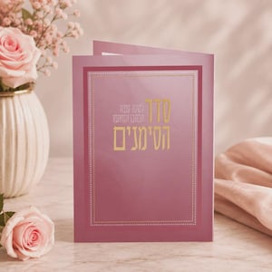 May include: A rectangular greeting card in shades of mauve and pink featuring gold Hebrew text centered on the front. The card is framed by a thin gold border with a dotted pattern and is displayed upright on a surface next to pink roses.