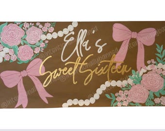 May include: Brown banner with the text "Ella's Sweet Sixteen" in gold and white script. The banner is decorated with pink bows, roses, pearls, and greenery. A festive decoration for a birthday celebration.