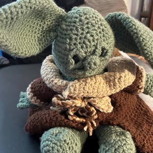 May include: Crocheted plush toy in the likeness of a character with large ears. The toy is primarily green, with a brown cloak and tan collar. The crochet stitches are visible, giving it a handmade appearance.