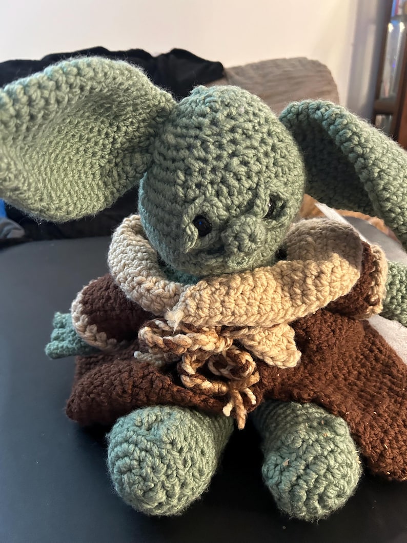 May include: Crocheted plush toy in the likeness of a character with large ears. The toy is primarily green, with a brown cloak and tan collar. The crochet stitches are visible, giving it a handmade appearance.