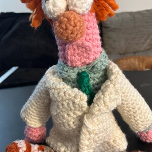 Pu&ograve; includere: Una bambola all'uncinetto fatta a mano di un personaggio con capelli arancioni, grandi occhi bianchi e un viso rosa. La bambola indossa un camice da laboratorio color crema, una cravatta verde e un colletto verde chiaro. I piedi della bambola sono arancioni e bianchi.