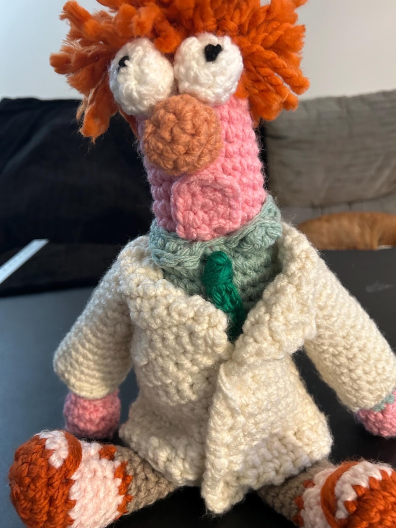 Pu&ograve; includere: Una bambola all'uncinetto fatta a mano di un personaggio con capelli arancioni, grandi occhi bianchi e un viso rosa. La bambola indossa un camice da laboratorio color crema, una cravatta verde e un colletto verde chiaro. I piedi della bambola sono arancioni e bianchi.