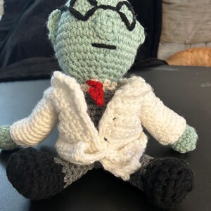 Pu&ograve; includere: Bambola all'uncinetto fatta a mano di uno scienziato. La bambola ha una testa verde chiaro con occhiali neri, una cravatta rossa e un camice bianco. Il corpo &egrave; grigio, con scarpe nere. La bambola &egrave; seduta.